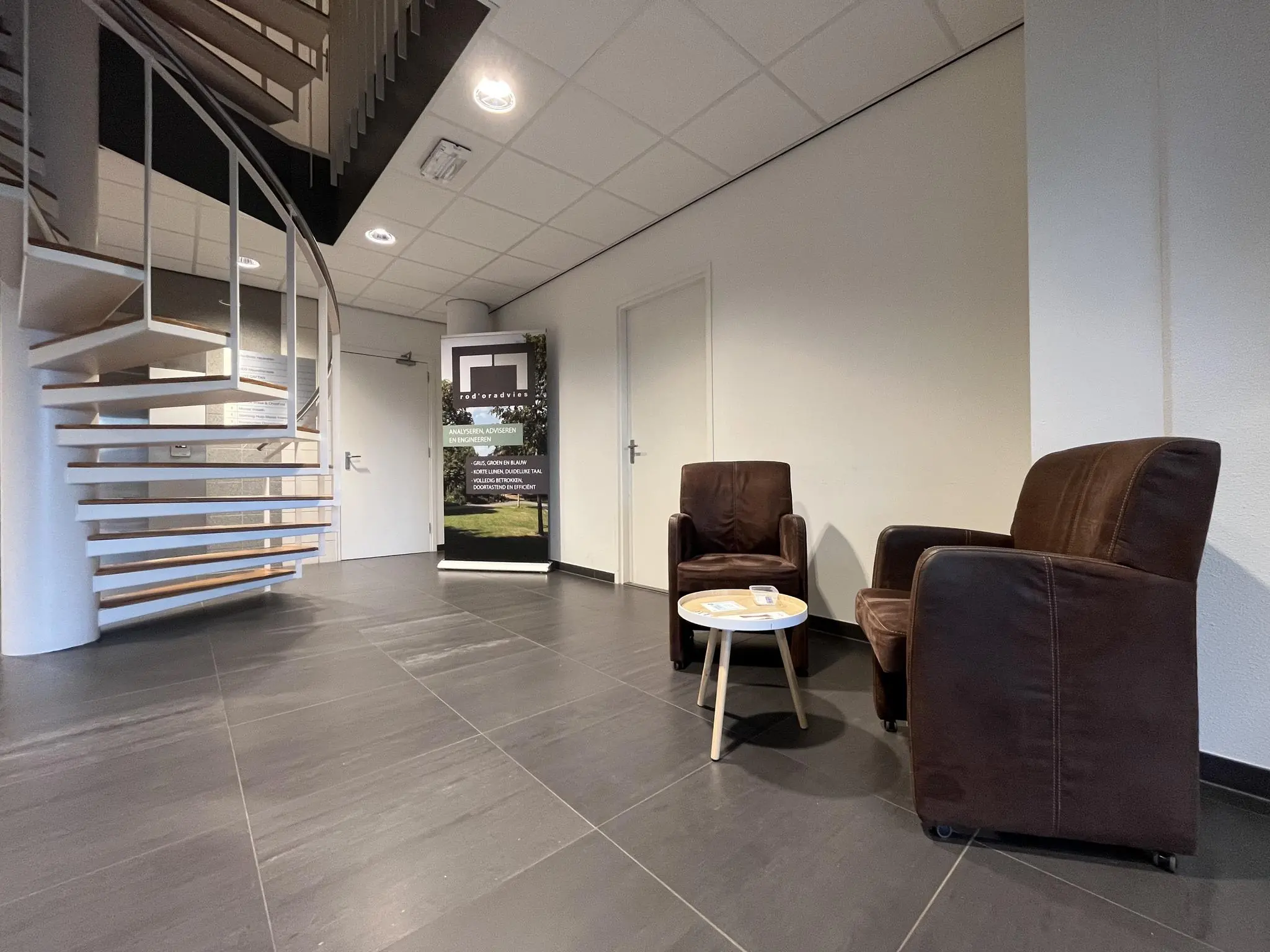 Wachtruimte met twee bruine fauteuils, een ronde tafel en een spiraaltrap in een kantoorpand in Leidse Rijn.